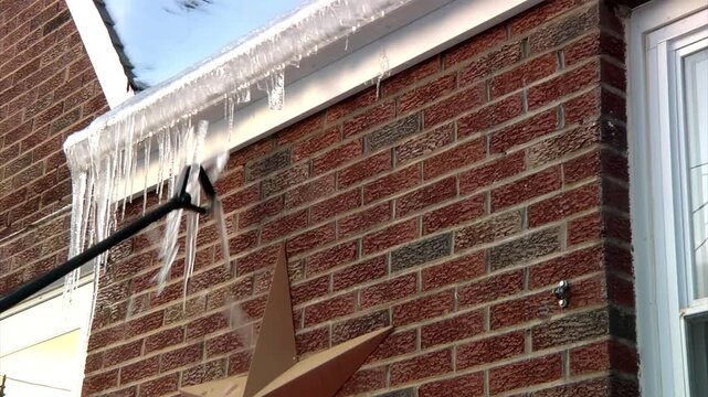 Homeowner uses a long tool to knock dangerous icicles off roof gutters, helping prevent ice dams and reduce winter storm damage to the house.