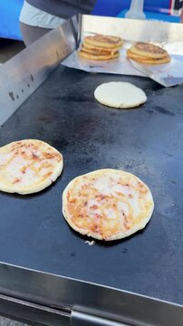 Pupusa flipping, cooking with spatula, grilled pupusas, Salvadoran food, flat-top griddle, golden patties, ethnic cuisine, outdoor vendor, stacked pupusas, Latin American snack, food in motion
