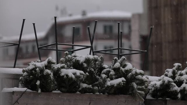 Snow blankets evergreen branches in wooden planters during a light snowfall in Cortina. Metal barstool frames stand inverted against a blurred backdrop of alpine buildings under a grey winter sky.