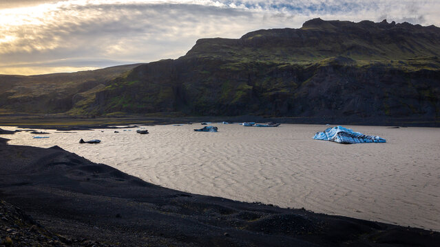Solheimajokull glacier lagoon with floating blue icebergs and volcanic ash moraines at sunset, Iceland