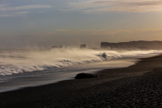 Dangerous sneaker waves crashing on Reynisfjara Black Sand Beach at sunset with Dyrholaey in the distance, Iceland