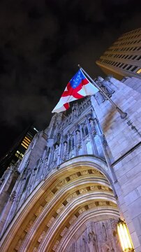 Approaching hand-carved stone fa&ccedil;ade of St. Tomas Church in New York, USA at night. Low angle view at the Episcopal Church flag flapping in the wind. Vertical video.