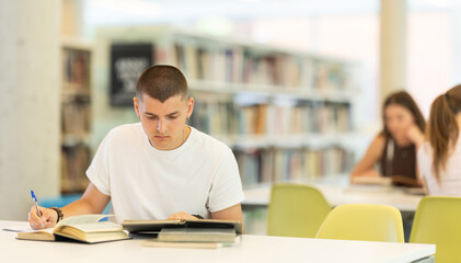 Guy reading books at table and writing notes in a notebook in a university library - preparing for exams