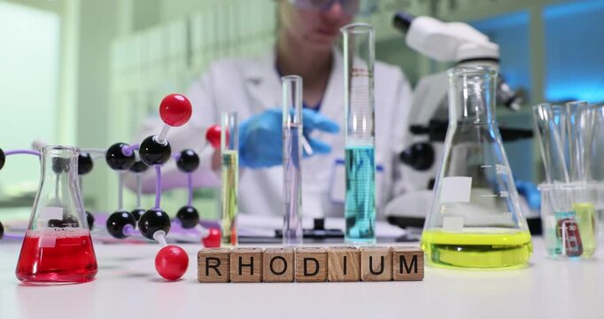Wooden cubes spell word Rhodium on laboratory table. Scientist woman works with colorful flasks near molecular model among glassware during experiment