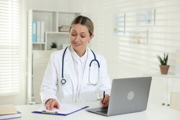 Smiling cardiologist with laptop, pen and clipboard working at table in clinic