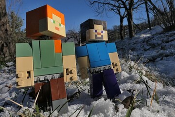 Naklejka premium Minecraft action figures of smiling Steve and Alex on a walk in winter garden, first snow visible on the lawn. Naked fruit trees and shrubs as well as clear blue skies in background. Afternoon sun. 