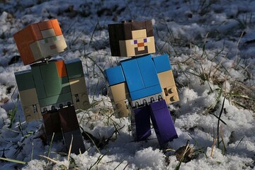 Naklejka premium Minecraft action figures of Steve and Alex walking on garden lawn covered with first winter snow, partially sunlit by afternoon sunshine, partially hidden in shadows of naked tree branches. 