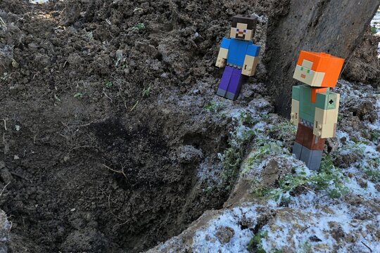Minecraft action figures of smiling Steve and Alex looking into deep dug out garden pit in winter half frozen garden soil. Traces of frost and snow visible on the edge. 