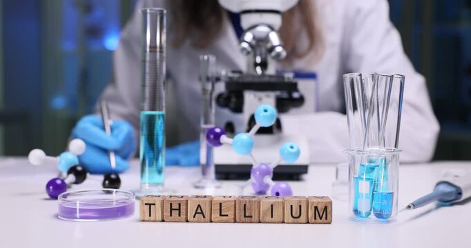 Wooden cubes spell word Thallium on laboratory table with glassware. Female doctor studies blue solution and colorful molecular models near microscope