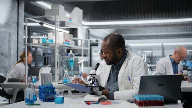 Lab researcher looking through scientific microscope lens to study cells. African american man performing biological sample examination with high tech optical equipment, camera A