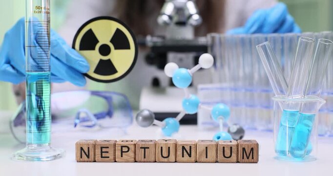 Wooden cubes spell word Neptunium near radioactive warning sign. Female doctor handles blue solution and test tubes in secure nuclear laboratory