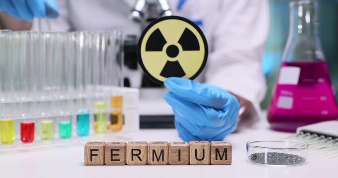 Wooden cubes spell word Fermium near radioactive warning label. Female doctor works with colorful solutions organizing test tubes in research lab