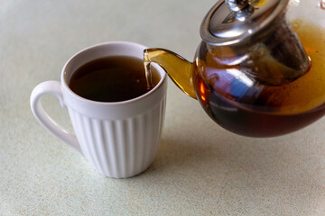 Close-up of hot tea being poured from a transparent glass teapot into a white ceramic mug on a clean, minimalist kitchen surface. Warm tones, natural light, and a calm atmosphere convey comfort, relax