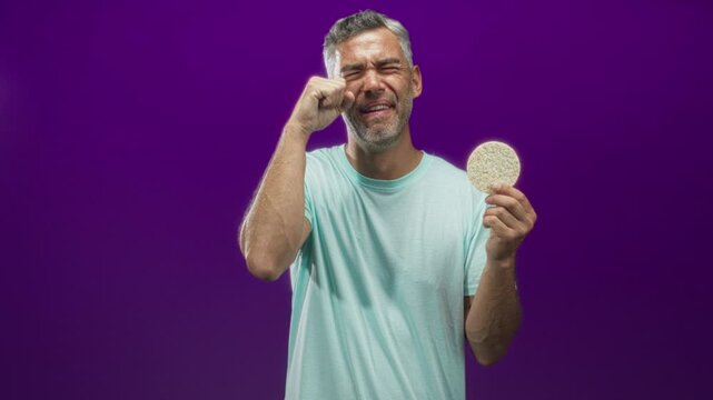 Man holding a round corn pastry in one hand and pinching his nose with the other in a purple studio set, face cringing at the smell; disgust.