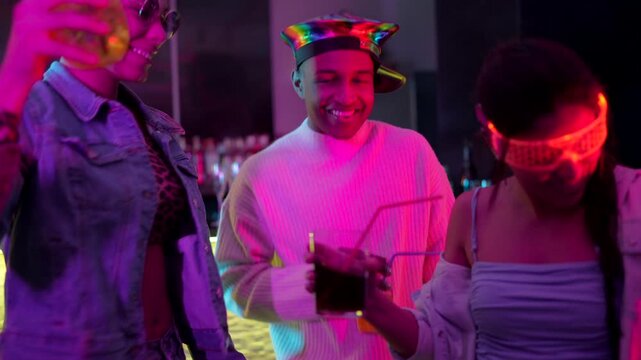 Three happy multiethnic friends wearing futuristic outfits drinking cocktails and dancing together at a bar with colorful neon lights, enjoying the nightlife and celebrating with drinks