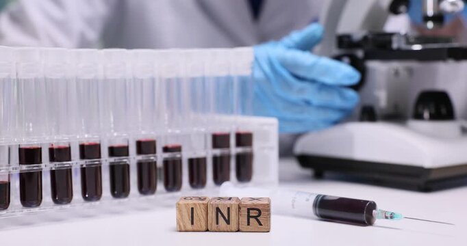 Wooden cubes spell abbreviation INR near blood filled test tubes. Laboratory worker monitors coagulation analysis adjusting microscope during test
