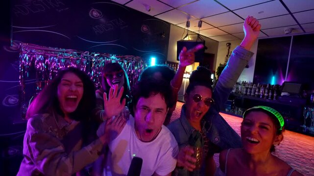 Happy young man singing into a microphone during a karaoke night, with his cheerful, diverse group of friends dancing, laughing, and singing along in a vibrant, neon lit bar setting