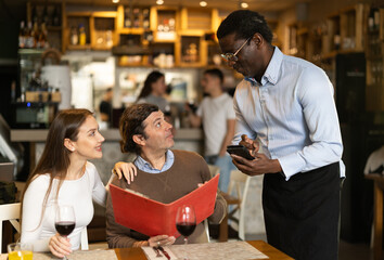 Obraz premium During communicating with couple customers, African man waiter writes down order of dishes and drinks, answers customers questions about offer from chef. Friends in restaurant makes order to waiter