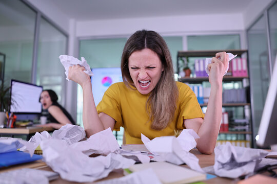 A young woman in a yellow shirt expresses her frustration among a clutter of crumpled papers. Behind her, a colleague reacts in surprise, capturing the chaos of a hectic workday.