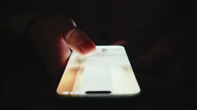 Extreme close-up male finger scrolling smartphone screen in deep darkness at night. Swiping finger, motion blur on phone screen. Evening online browsing, digital content consumption