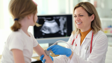 Obraz premium A kind doctor smiles warmly as she examines a young girl's arm with a medical device in a well-lit clinic. The atmosphere is friendly and reassuring, promoting health and care.