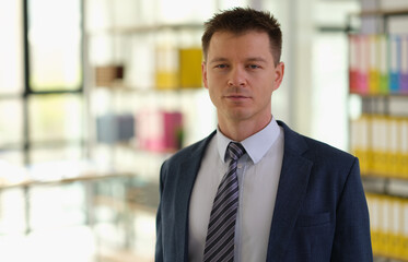 Fototapeta premium A professional man in a sharp suit poses confidently in a sleek workspace. Bright sunlight filters through large windows, highlighting organized shelves of colorful documentation behind him.