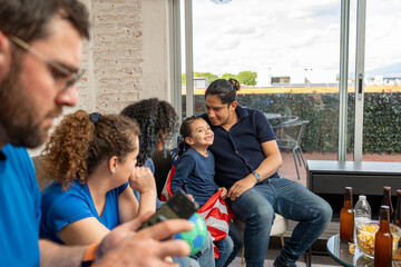 Obraz premium Girl wearing a USA flag looks at the camera while family and friends enjoy watching football soccer at home. Sports togetherness, cultural pride, joyful moments, friendship, casual home viewing