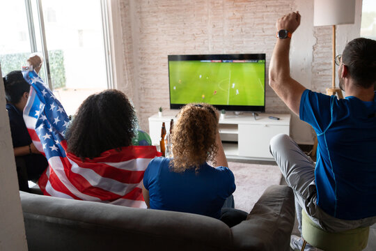 Group celebrating together with a USA flag as they watch football soccer on TV at home. Sports victory mood, friendship, national pride, emotional connection, casual indoor gathering