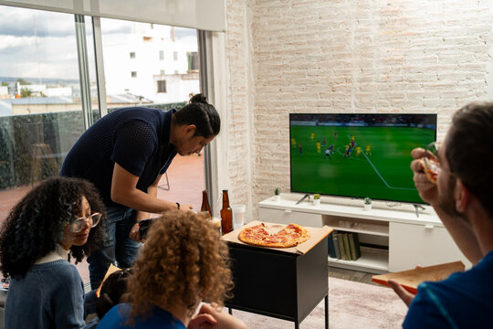 Friends eating pizza while watching football soccer on TV, the screen clearly visible during the match. Sports snacks, casual gathering, friendship, shared enjoyment, home viewing