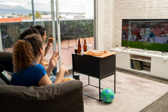 Group of friends focused on a TV screen watching football soccer while sharing pizza at home. Sports anticipation, social connection, friendship, casual indoor gathering, match atmosphere