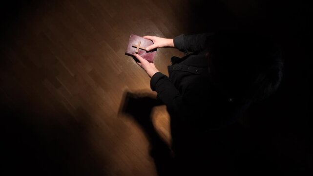 Man in dark clothing holding a bible with a wooden cross, finding comfort and hope in prayer and faith during a moment of spiritual reflection and introspection
