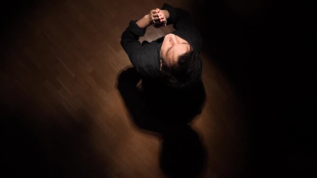 Depressed man looking up with clasped hands, seeking comfort and spiritual guidance, expressing faith and inner reflection in a dramatic moody light with shadow on a wooden floor