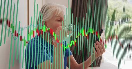 Obraz premium Holding clear mug, senior woman wearing blue top checking phone at home, with stock chart overlay