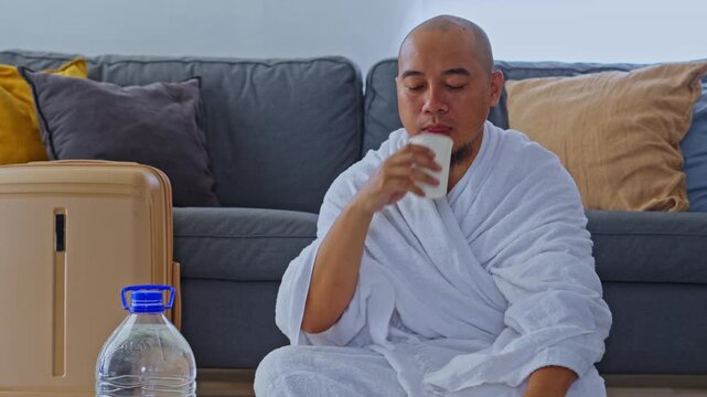 Muslim pilgrim in Ihram finishing dua and drinking zamzam water from cup while sitting on floor