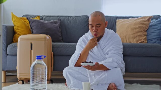 Muslim pilgrim in Ihram eating dates near suitcase and zamzam water while sitting on floor