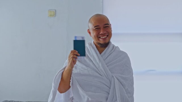 Cheerful pilgrim in ihram attire showing travel document and giving thumbs up before Hajj or Umroh