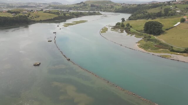 Aerial drone footage of the Suances estuary looking inland, showing the Saja-Besaya river with its deeper blue channel bordered by stone groynes and riprap; green meadows can be seen