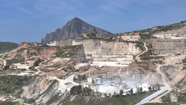 AERIAL: Active marble quarry with terraced white cuts and a processing plant sending dust into the air. Rugged hills and a mountain peak rise behind the industrial site in western part of Sicily.