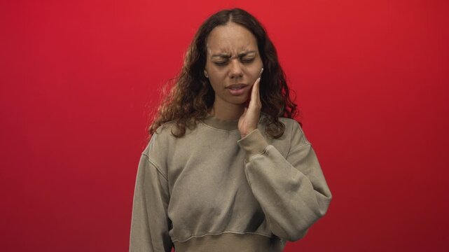 Young woman holding cheek with hand, eyes closed and wincing for toothache in studio against red wall; pain dental concern.
