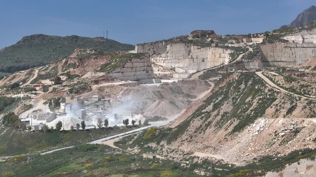 AERIAL: Stepped walls of a marble quarry and stone processing plant, where white dust hangs over conveyor belts and stockpiles. Industrial extraction in the hills of the Trapani Province of Sicily.