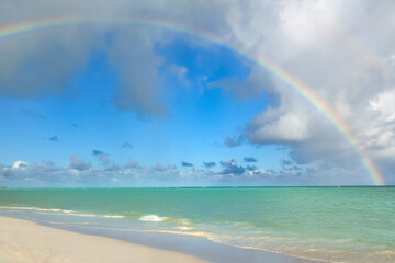 Scenic tropical landscape, peaceful turquoise waters of the sea, white sand and the cloudy sky with a wonderful bright rainbow.