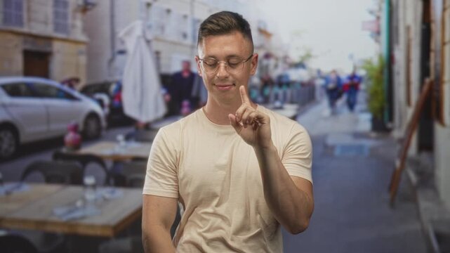 Young man with glasses touches chest and raises index finger beside cafe table on a narrow street; calm reflection.
