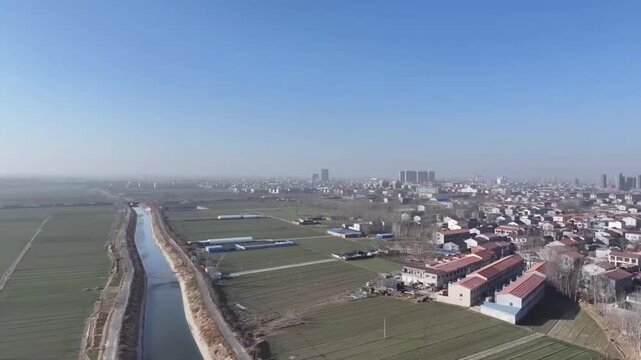 A serene 1080p aerial footage captured by a drone flying over the vibrant rural landscape of China. The left side of the frame showcases vast, cultivated farmland, while on the right, a cluster of tra