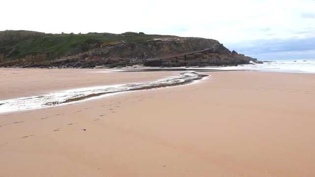 Wide sandy beach at Praia das Ma&ccedil;&atilde;s with a small river meeting the Atlantic, rocky shoreline, and calm cloudy atmosphere.