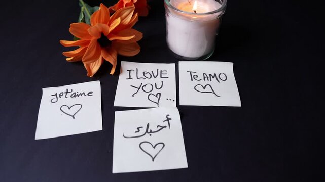 Overhead close-up of a lit jar candle, orange flowers, and sticky notes saying &ldquo;I love you&rdquo; in multiple languages on a dark tabletop, creating a warm romantic indoor mood.