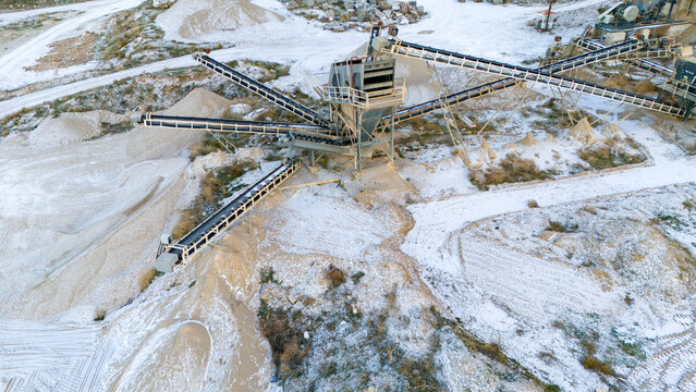 Wide aerial drone perspective of an active mining quarry facility in winter. Heavy industrial machinery and long conveyor belts process raw aggregate materials over the snowy ground.