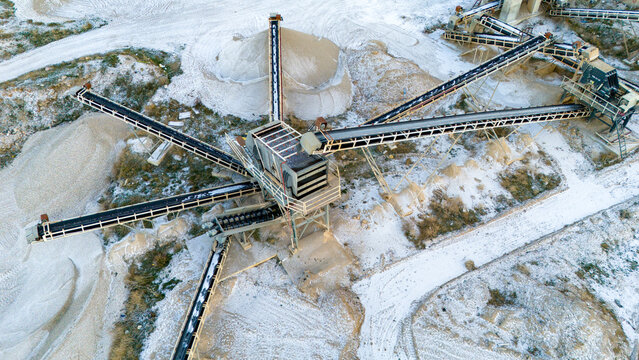Top-down aerial drone view of radial conveyor belts extending from a central stone crushing machine. The industrial quarry site is covered in a layer of fresh white winter snow.