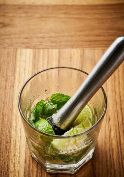 Close-up of lime, mint, and sugar being muddled in a glass on a wooden table, preparing a refreshing cocktail.