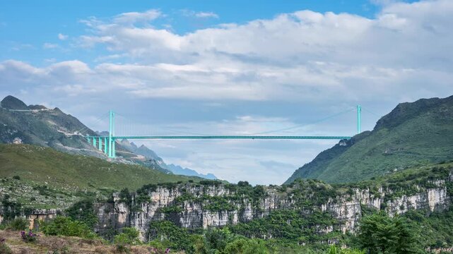 Time-lapse of the Huajiang Canyon Bridge under a bright blue sky with fluffy clouds, spanning a dramatic mountain gorge in Guizhou Province, China.