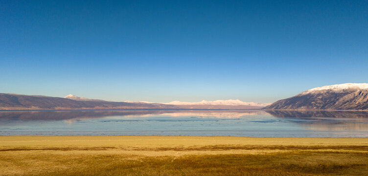 Ground level shot of a vast dry field leading to a clear blue lake under a bright afternoon sky with a snow capped mountain in the distance